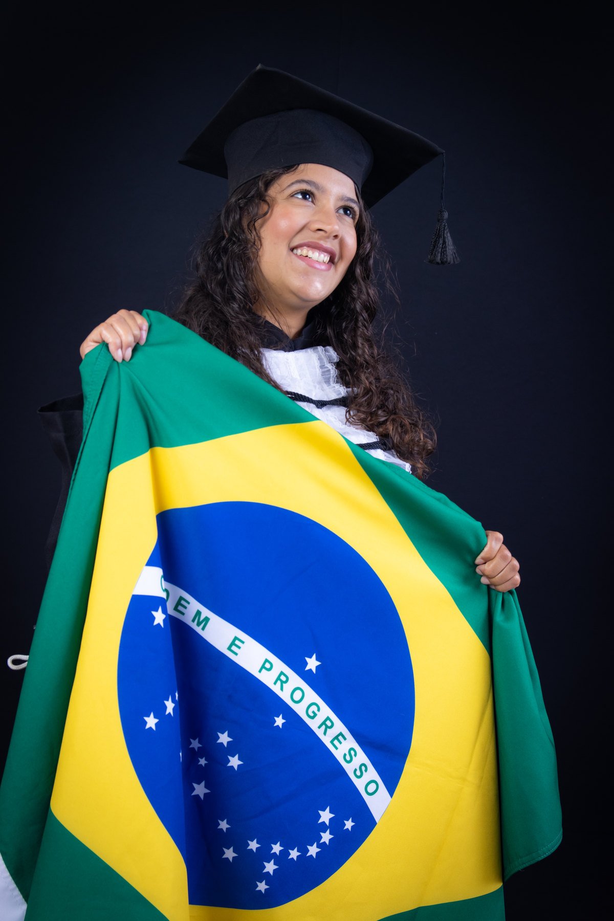 Formanda de beca e capelo sorrindo com a bandeira do Brasil aberta nas mãos, no fundo preto
