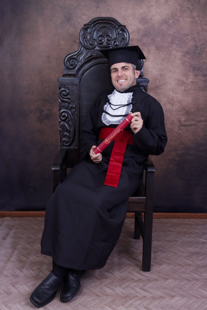 Formando de Odontologia de beca e faixa vermelha sentado na Cadeira Colonial Luiz XV com o canudo do curso