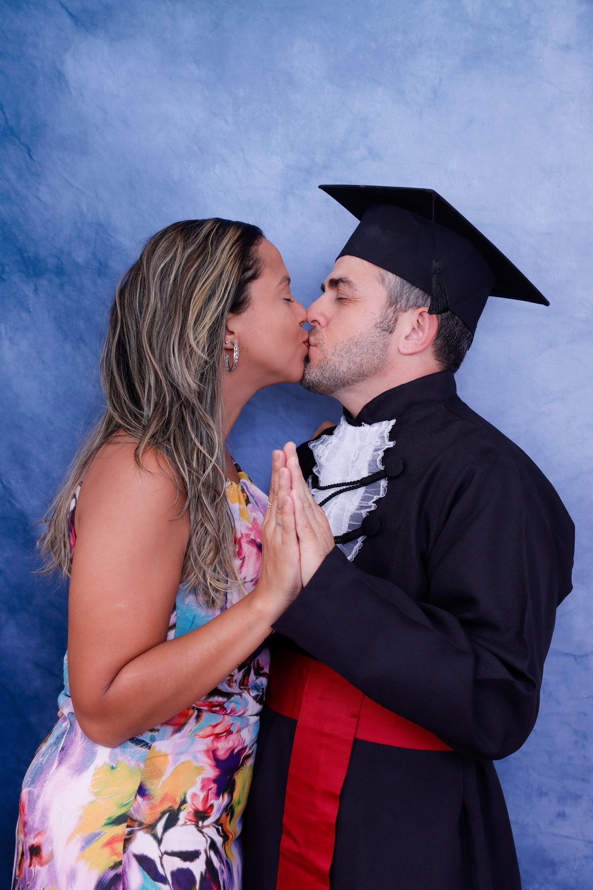 Formando de Odontologia de beca e capelo beijando a companheira no fundo azul