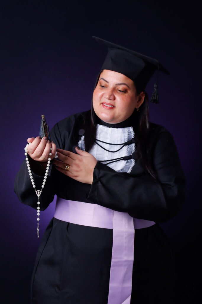 Formanda de Pedagogia de beca olhando para uma imagem de Nossa Senhora e um terço nas mãos