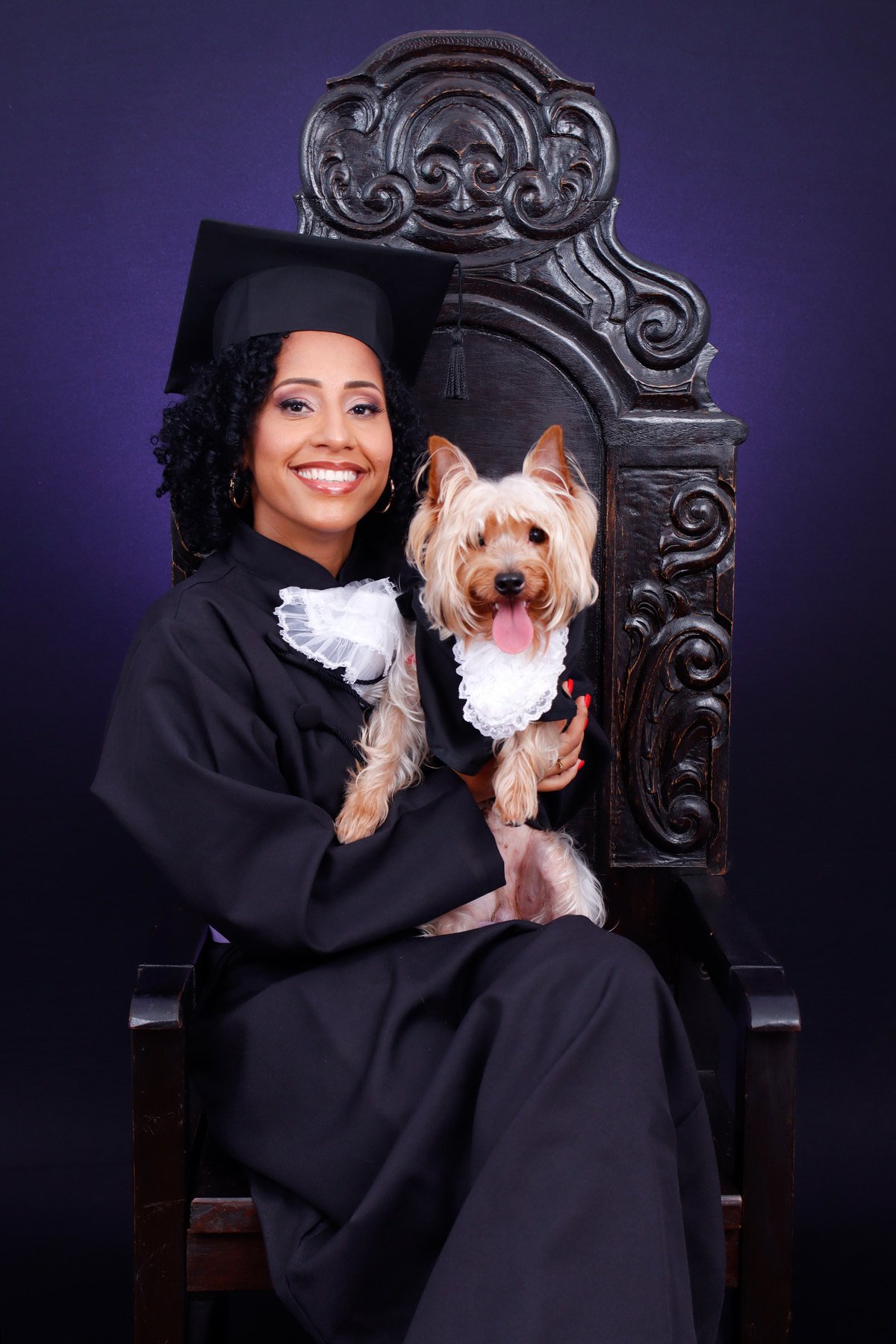 Formanda de beca e capelo na Cadeira Colonial Luiz XV com o cachorro yorkshire vestido de beca no colo