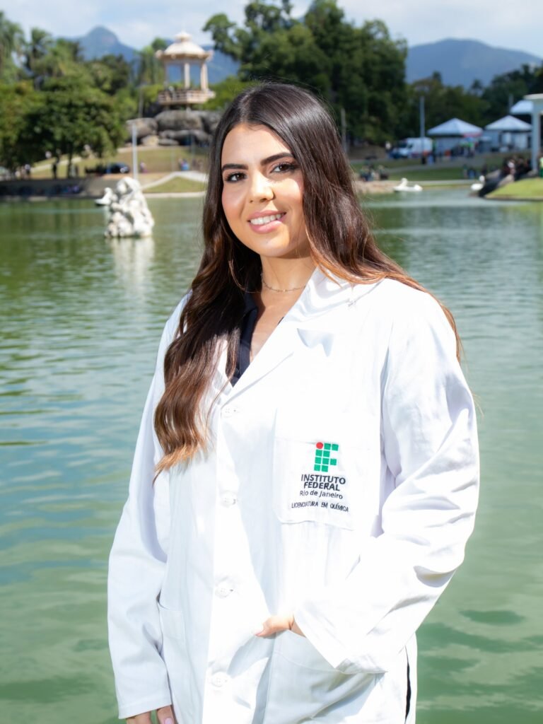 Formanda de Licenciatura em Química do IFRJ de jaleco bordado à beira do lago da Quinta da Boa Vista, com o coreto ao fundo