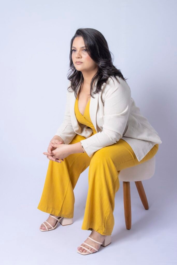 Maquiadora de macacão amarelo e blazer bege sentada em banqueta
