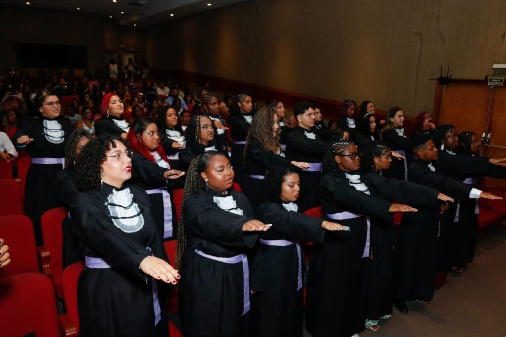 Turma de formandos com o braço estendido fazendo o juramento na colação de grau, em auditório