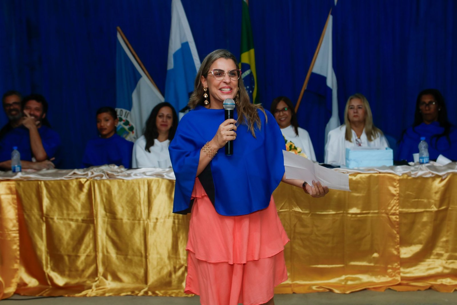 Paraninfa discursando ao microfone diante da mesa de honra na colação de grau