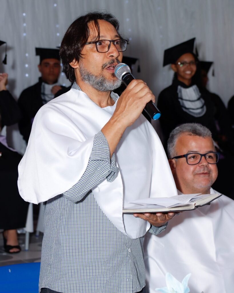Paraninfo lendo o discurso ao microfone, com os formandos de beca ao fundo